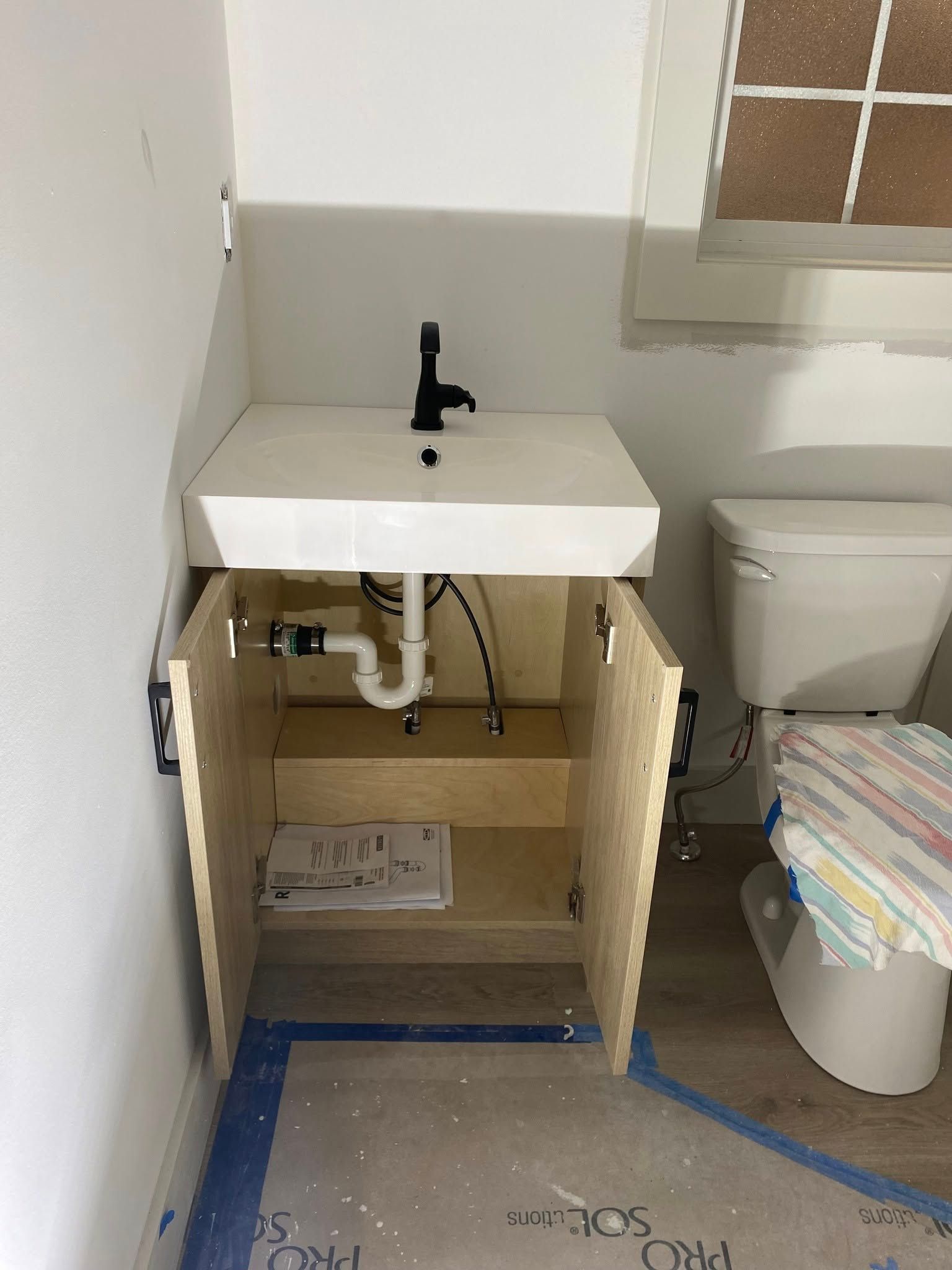 A wall-mounted bathroom vanity with an open cabinet, white sink, black faucet, and an exposed P-trap, next to a toilet.