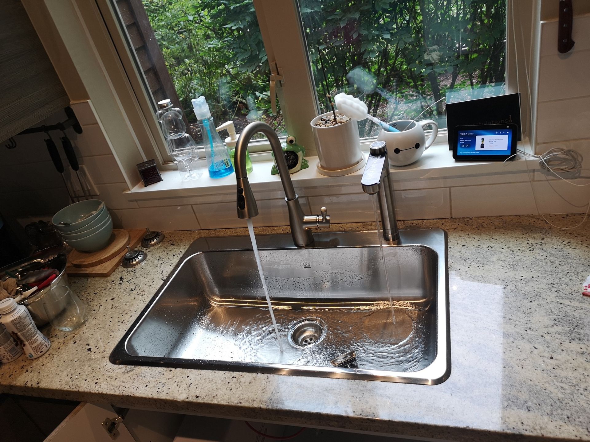 A stainless steel kitchen sink with water running from the faucet, set against a window with various items on the sill.