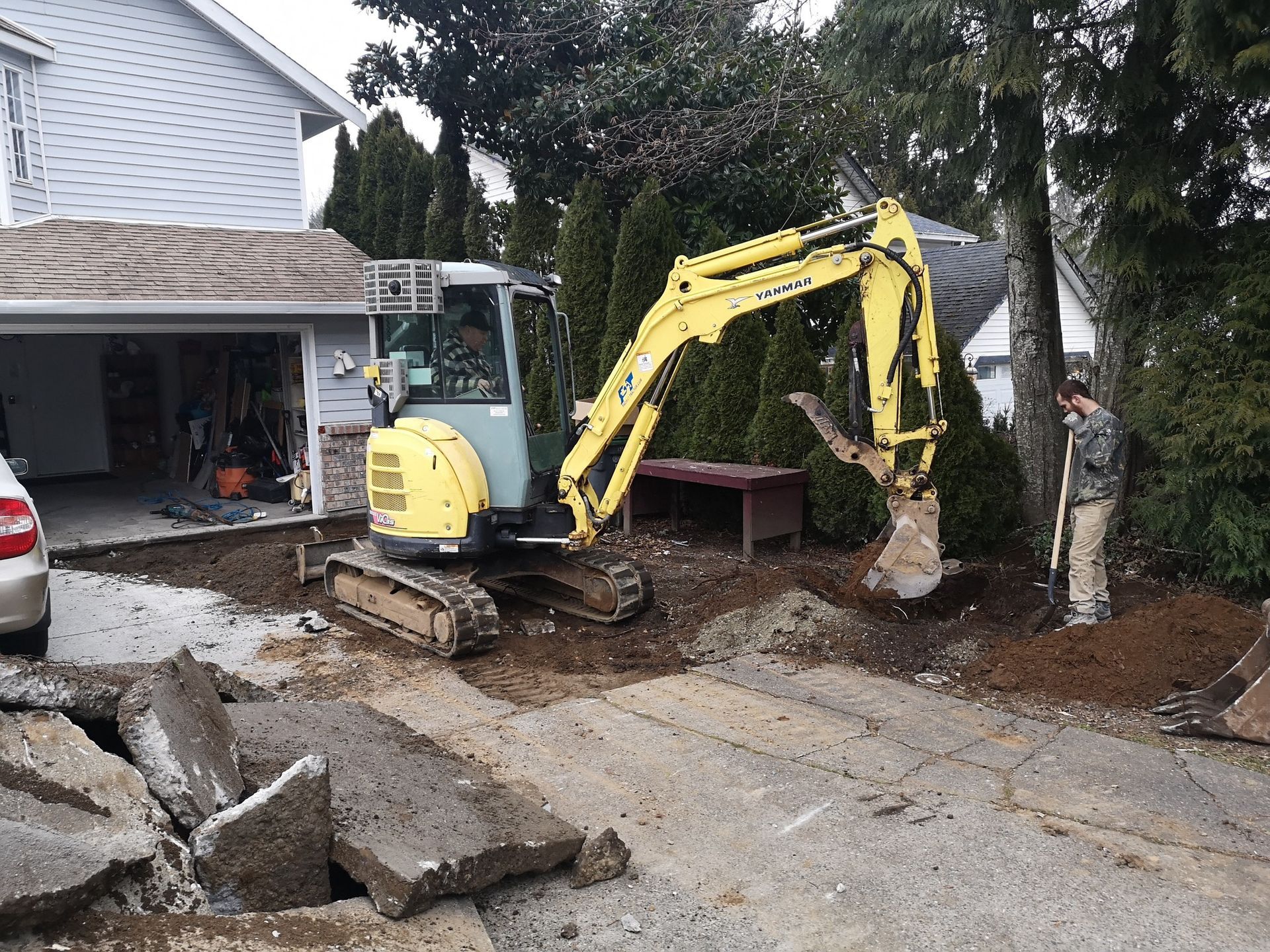 A yellow excavator is parked on a residential driveway, digging into soil near a garage while a person stands nearby.