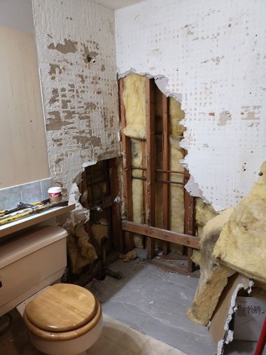 A bathroom undergoing renovation with damaged drywall exposing wooden wall studs, insulation, and plumbing.