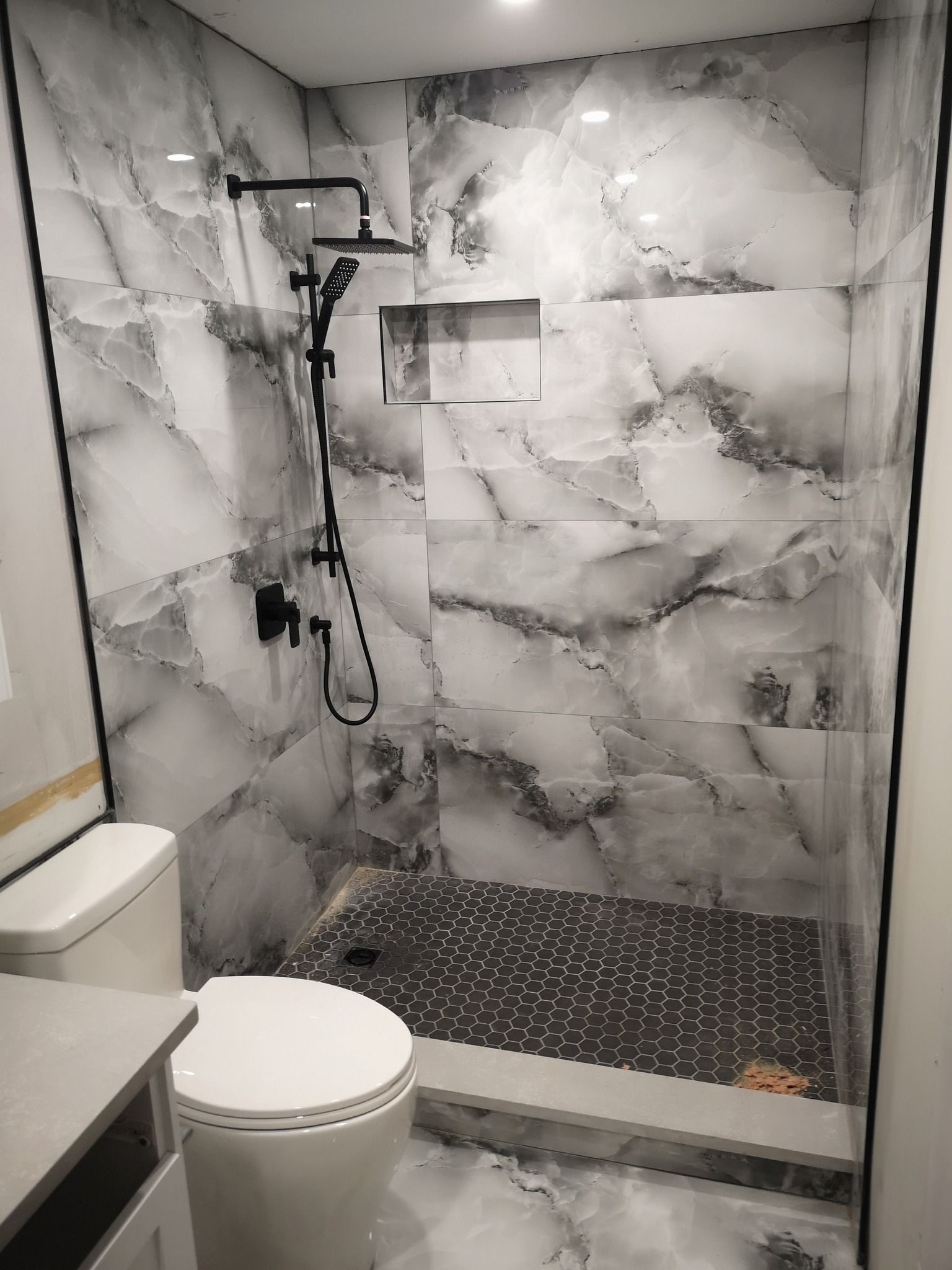 A small bathroom with a walk-in shower featuring white marble-patterned walls and a dark, patterned tiled shower floor.