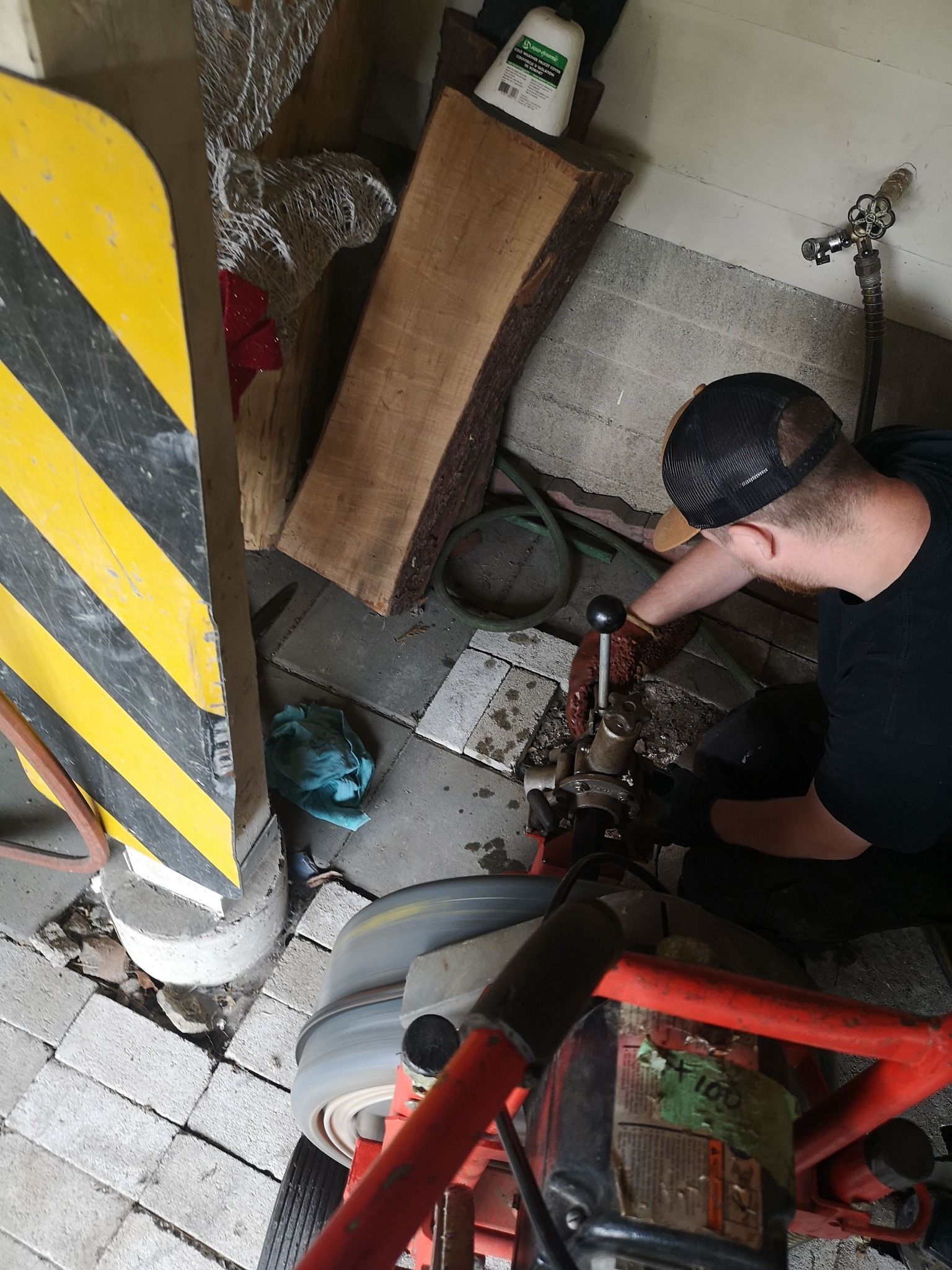 A person wearing a black cap and gloves operates a red industrial floor cleaner on a tiled floor in a garage area.