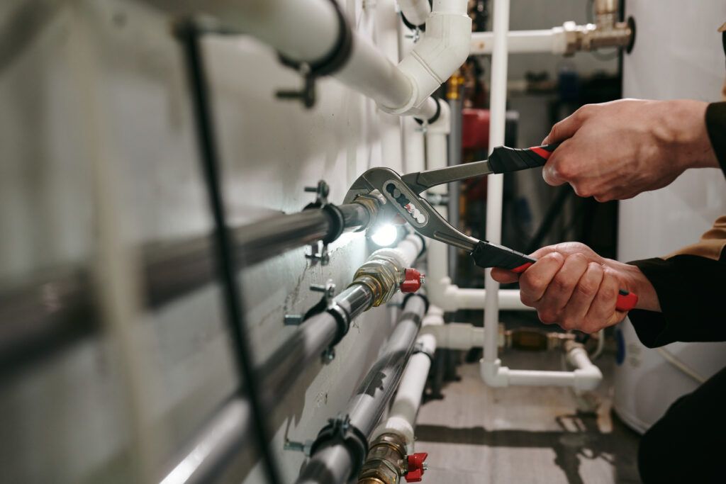 A person uses pliers to tighten a metal pipe fitting on a wall-mounted plumbing system in a utility room.