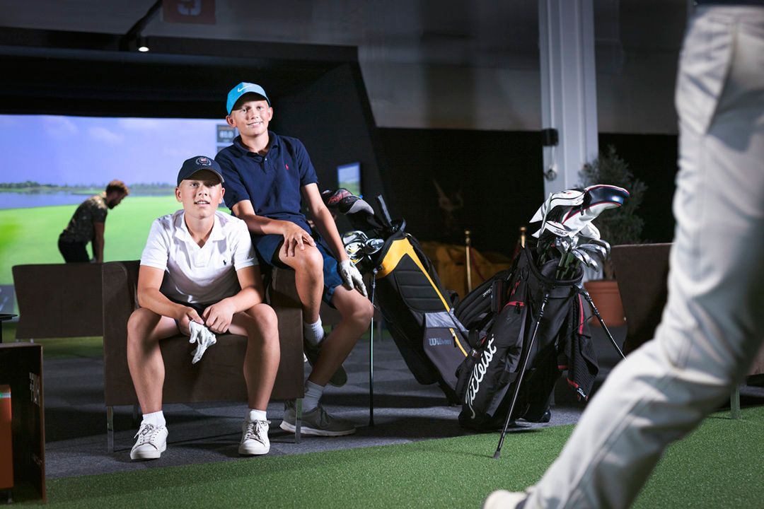 Two young men relaxing in an indoor golf setting; one sitting, one leaning. Golf clubs are nearby.
