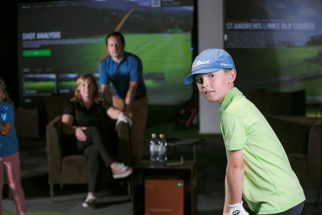 Boy in green shirt and blue cap at golf simulator, with parents watching.