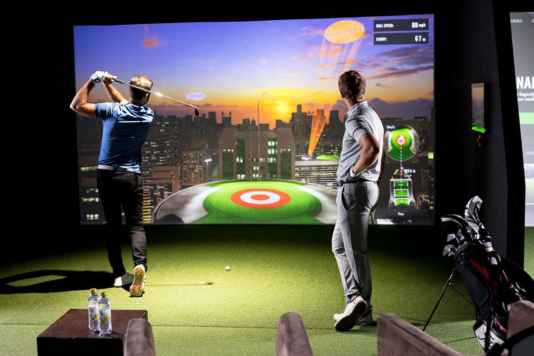 Two men playing golf in a virtual golf simulator with a city skyline backdrop.