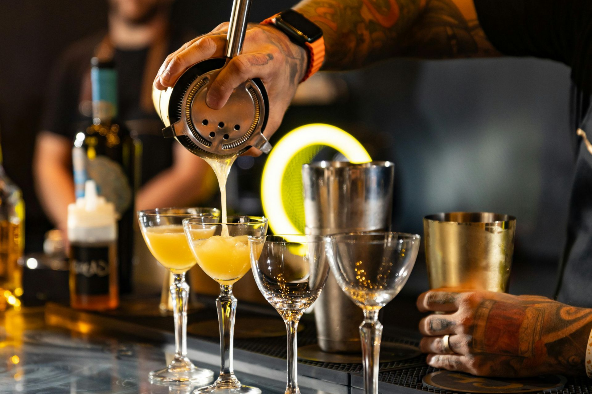 Bartender pouring cocktails into glasses on a bar. Yellow drink, tattoos visible.