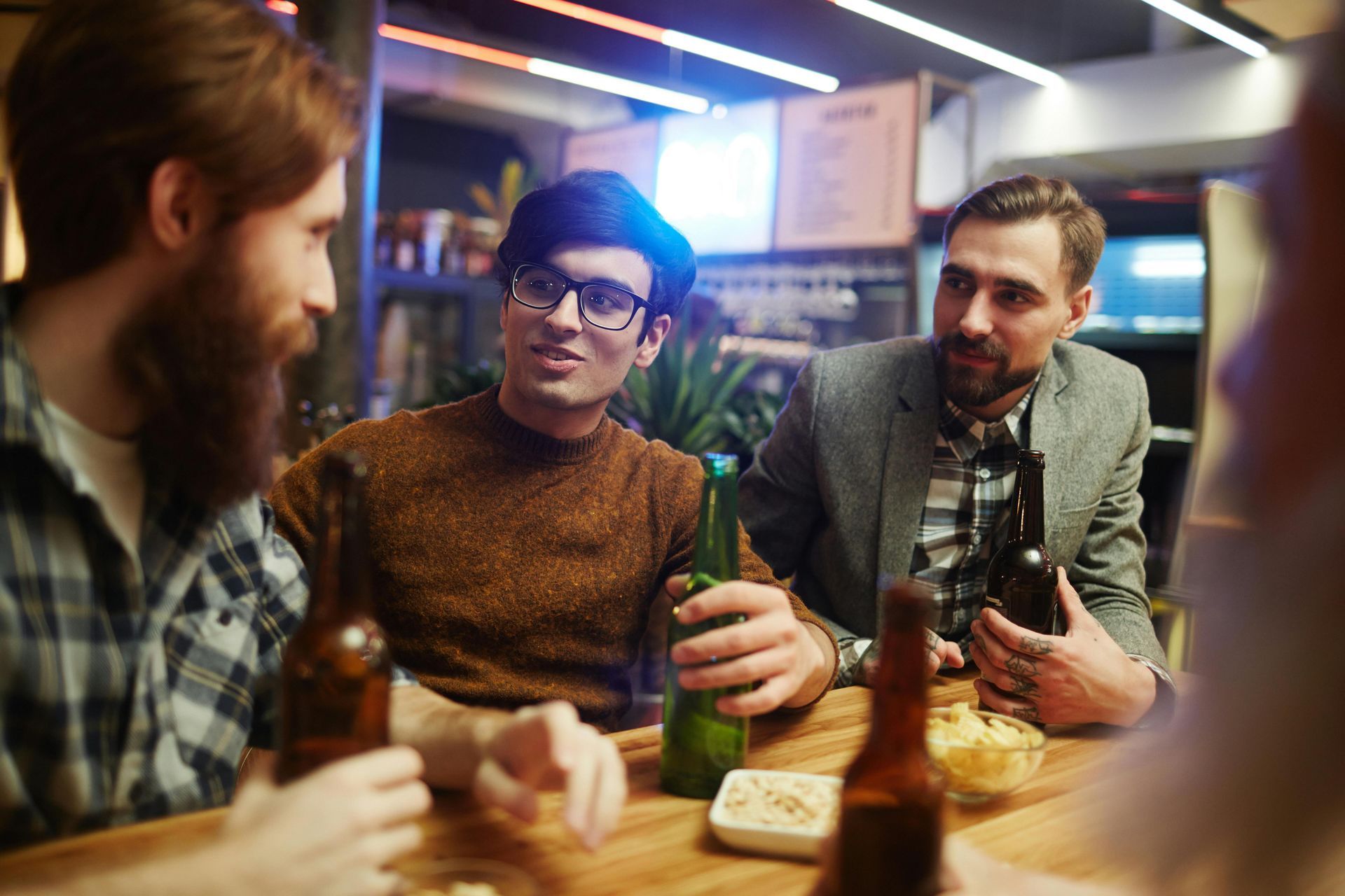 Three men seated at a bar, drinking beer and conversing. Interior setting with snacks.