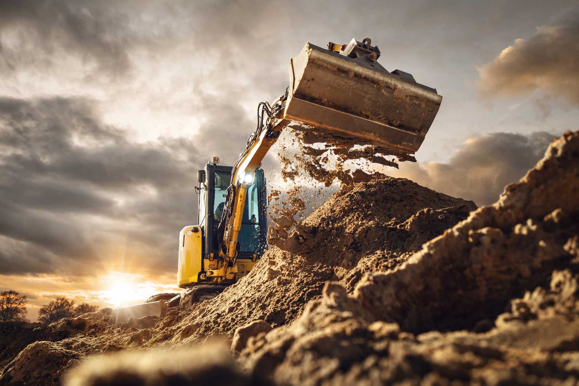 Yellow excavator digging dirt with a cloudy sky and sunset in the background.