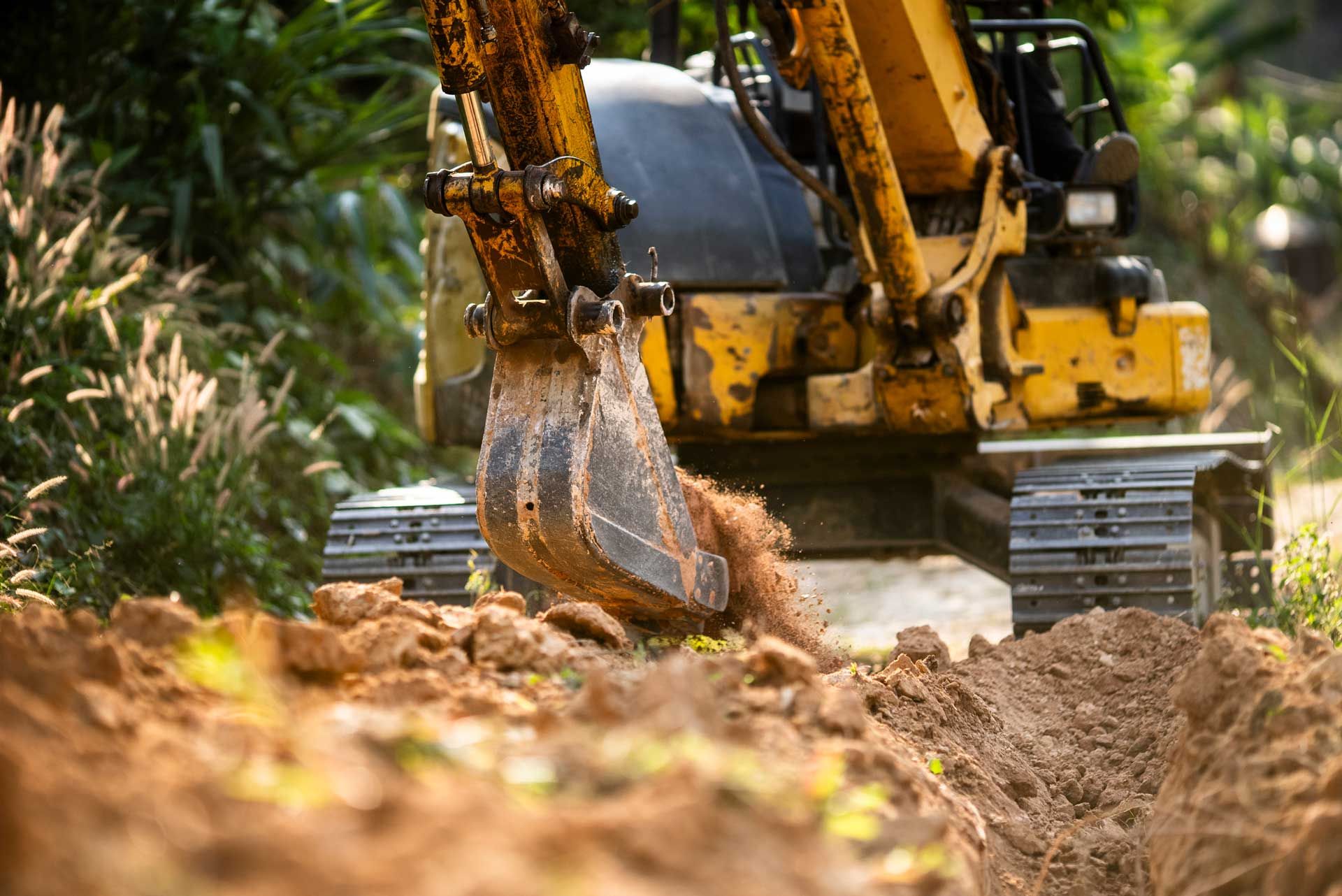Yellow excavator digging a trench in the dirt, surrounded by green foliage.