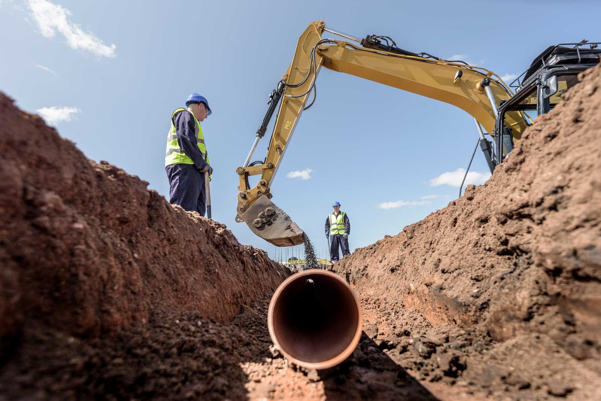 Excavator digging trench for brown pipe with workers in vests and hard hats on site.