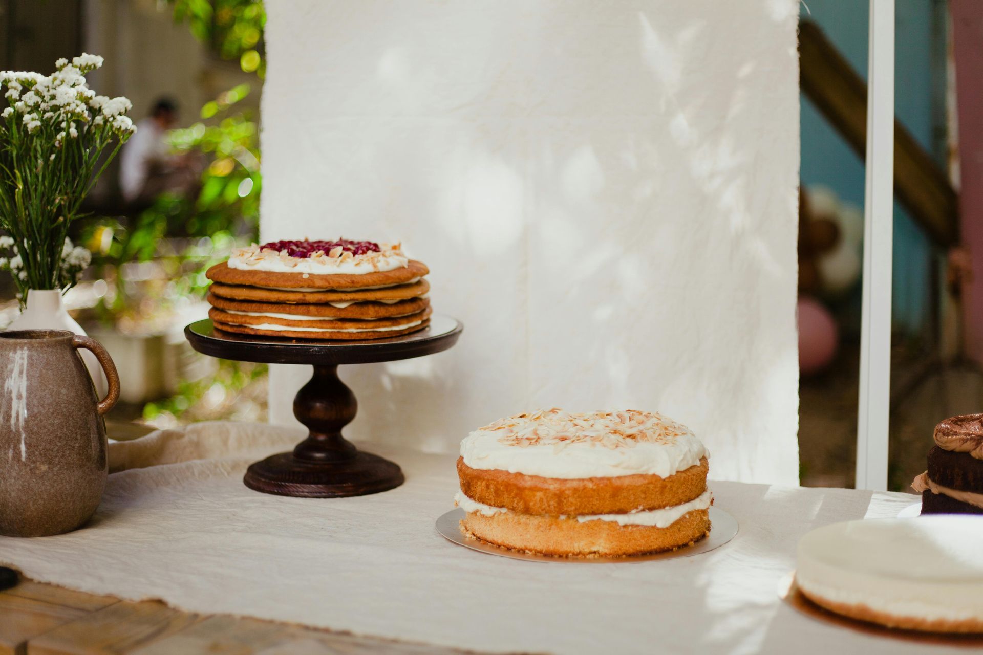 Two cakes, one layered with pomegranate seeds on a stand and one plain with frosting, sit on a table with flowers.