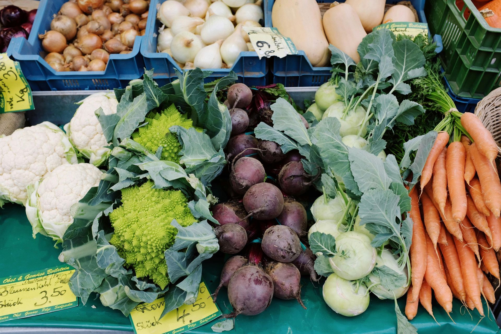 Fresh produce at a market, including cauliflower, romanesco, beets, kohlrabi, carrots, onions, and squash.