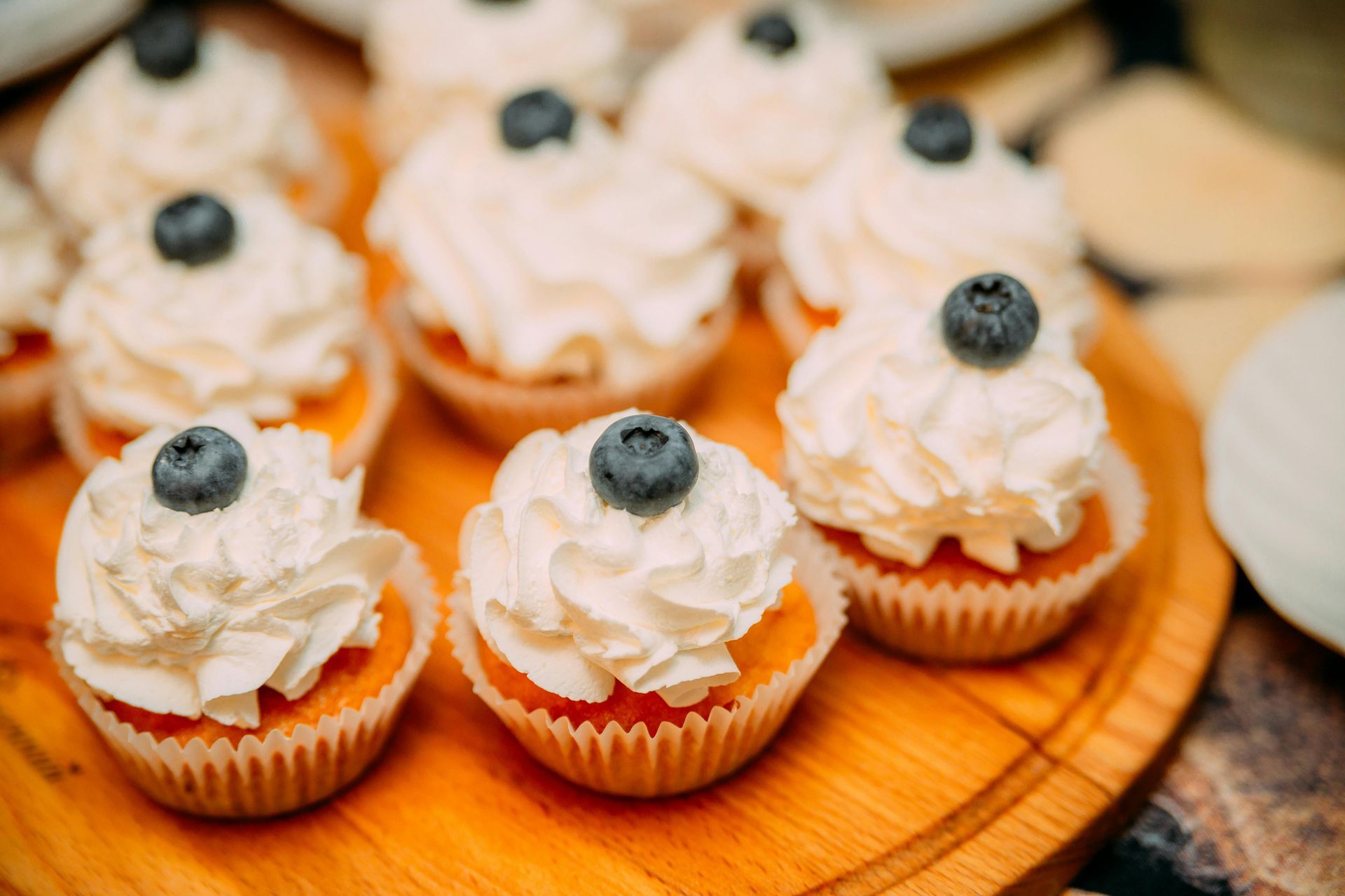 A group of small cupcakes topped with white frosting and a single fresh blueberry on a round wooden serving board.