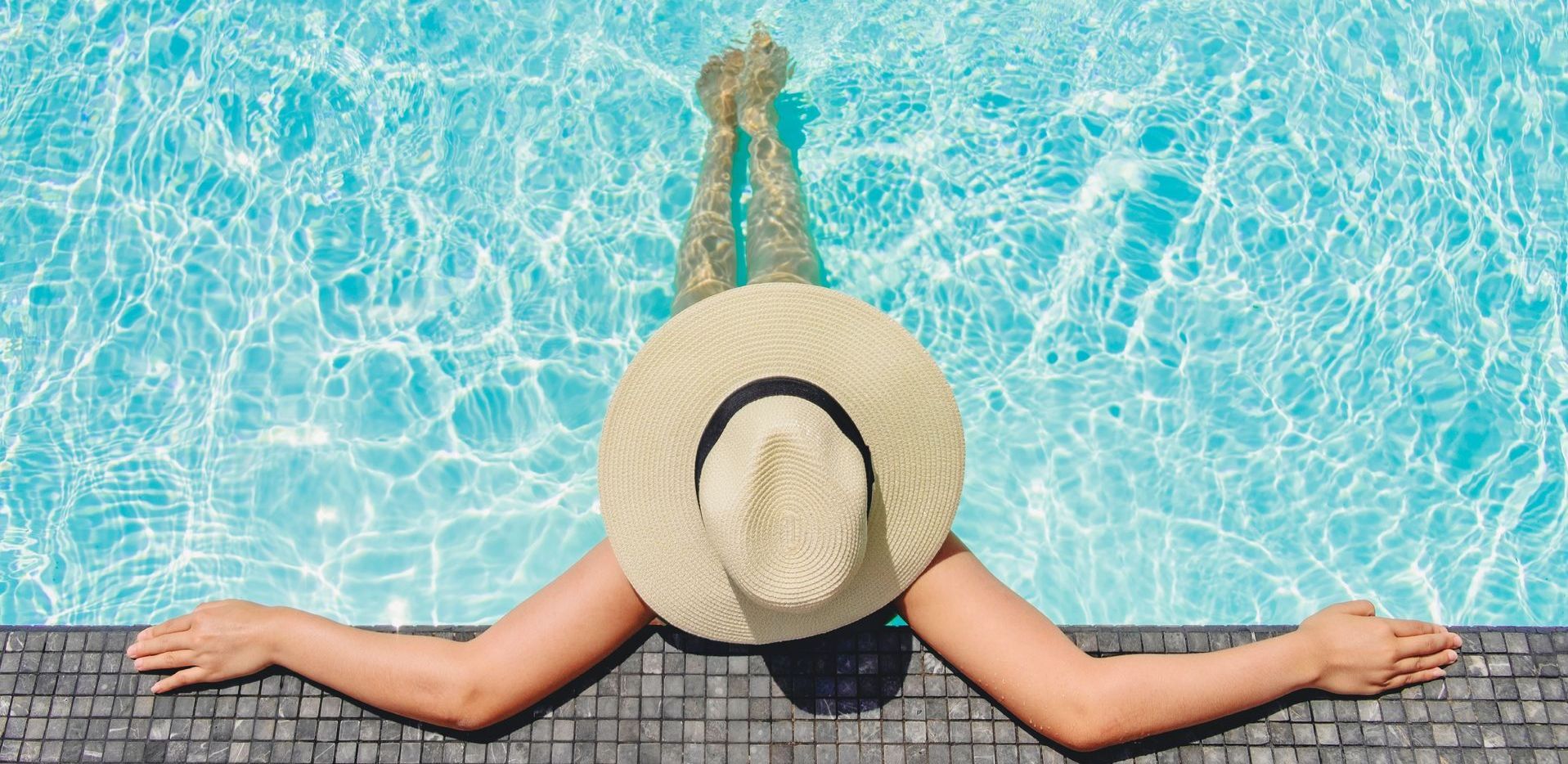 A woman in a hat is laying on the edge of a swimming pool.