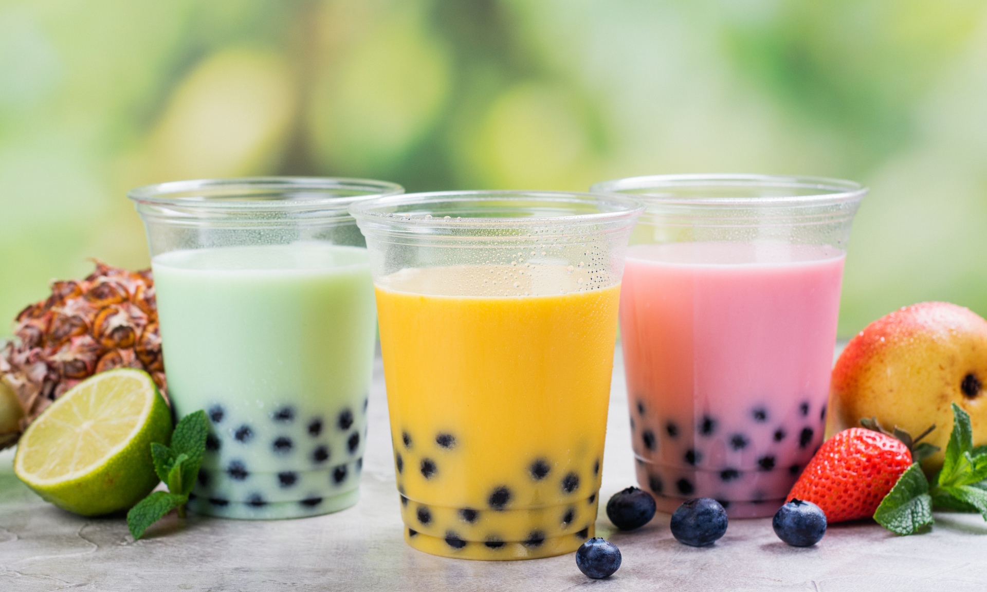Three colorful boba tea drinks: green, yellow, and pink. Fruit and mint garnish on a light surface.