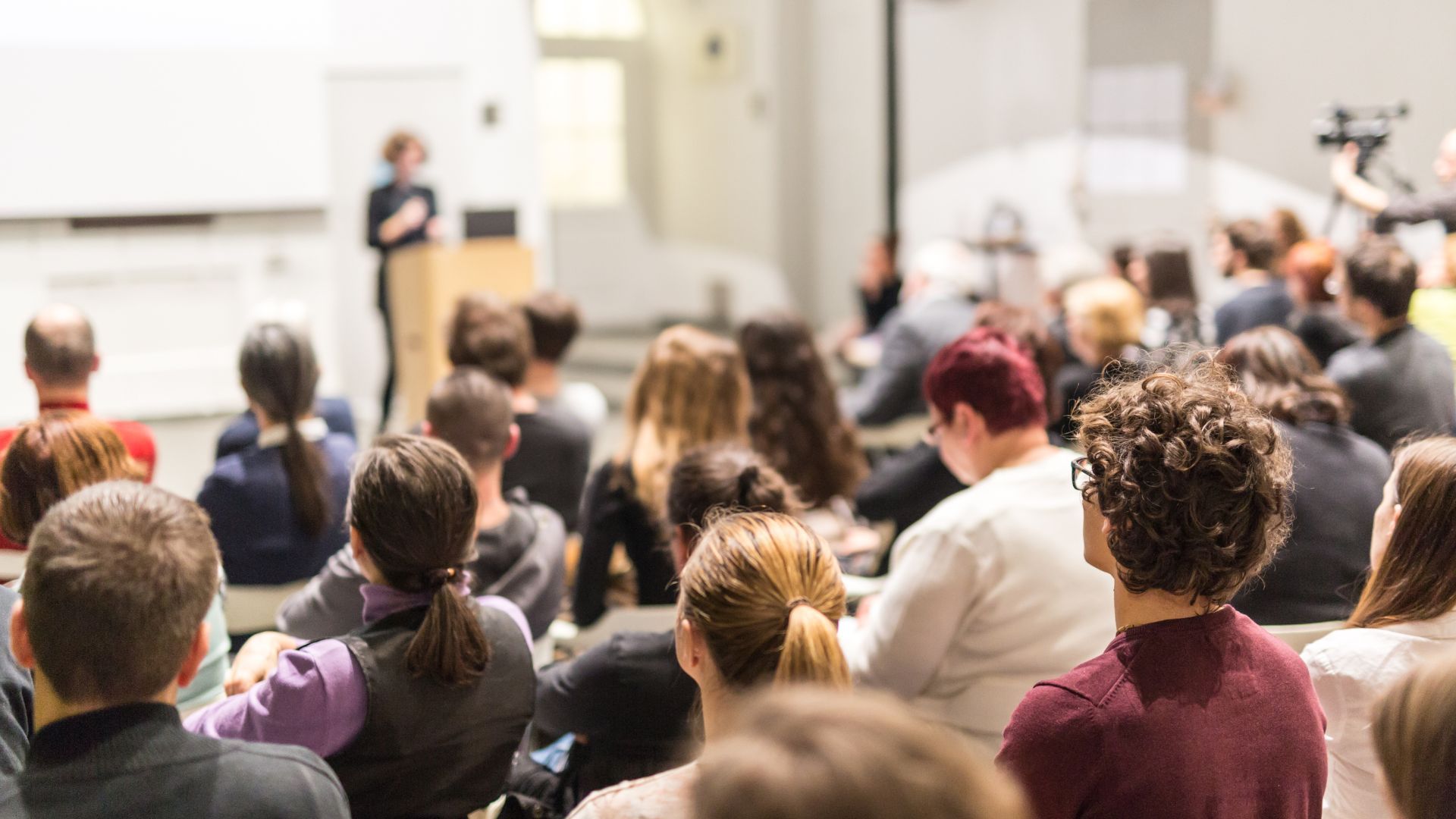 A large group of people are sitting in a lecture hall watching a presentation.