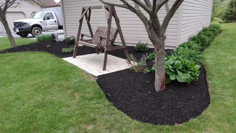 A wooden porch swing sits on a stone pad within a landscaped garden bed with dark mulch beside a tree and house.