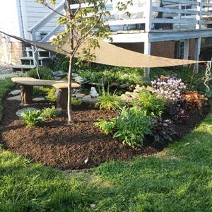 A tan shade sail hangs over a backyard garden bed featuring a curved bench, small tree, various plants, and stone accents.