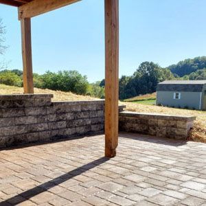 A stone paver patio with a low retaining wall under a wooden pergola, looking out toward a small shed and grassy hills.