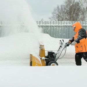A person in an orange winter jacket uses a yellow snow blower to clear a path on a snowy day.