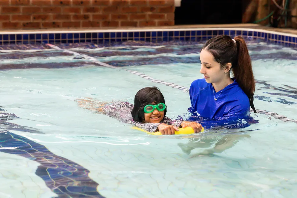 Child with goggles and a float swims with instructor in a pool.