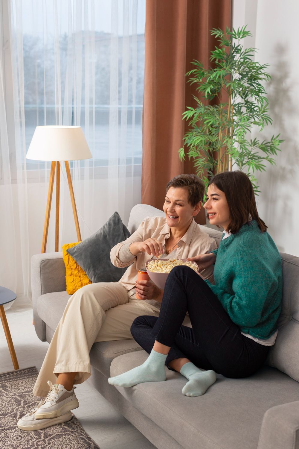 Two women are sitting on a couch eating popcorn and watching tv.