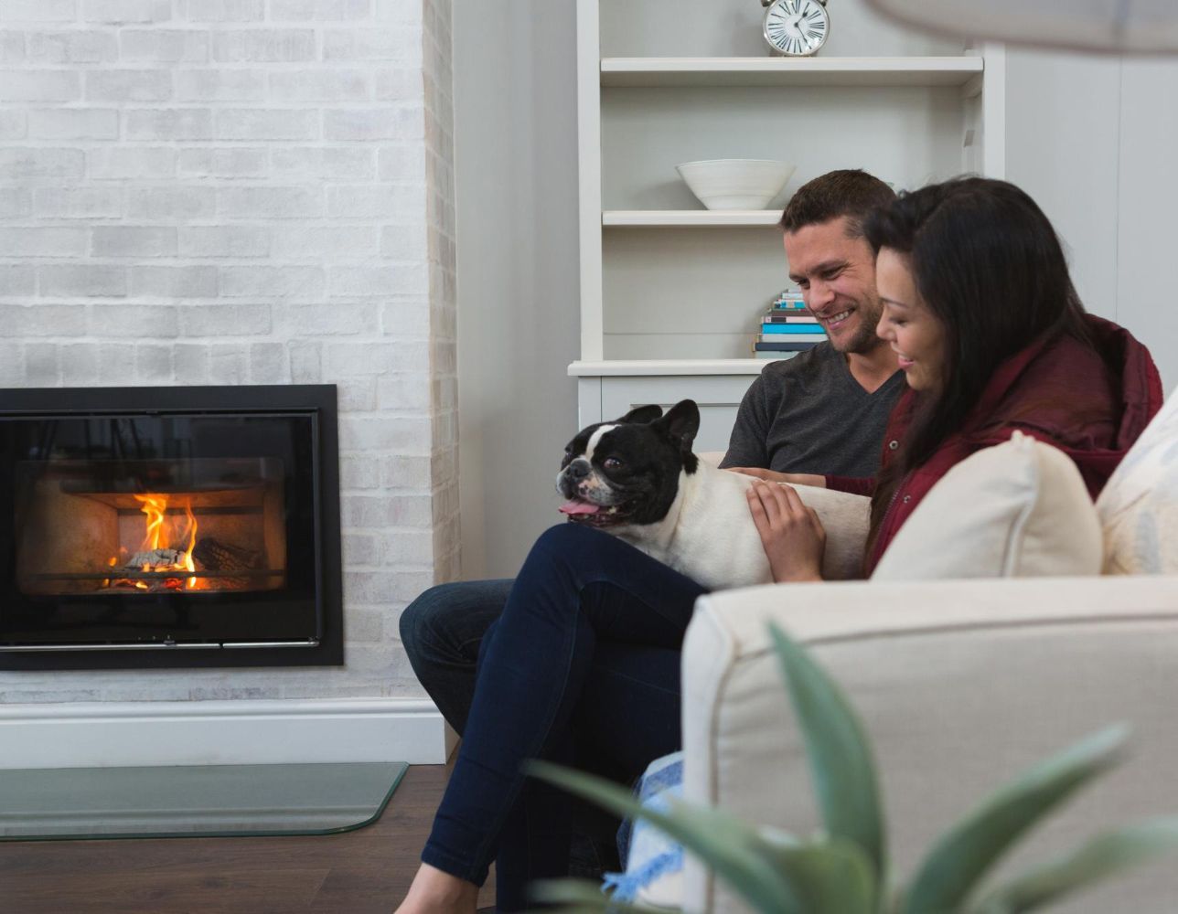 A man and woman are sitting on a couch with a dog in front of a fireplace.