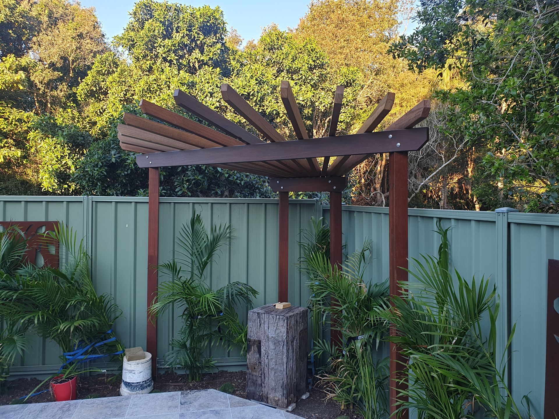 Wooden pergola with brown support posts and radiating slats; in a garden setting.