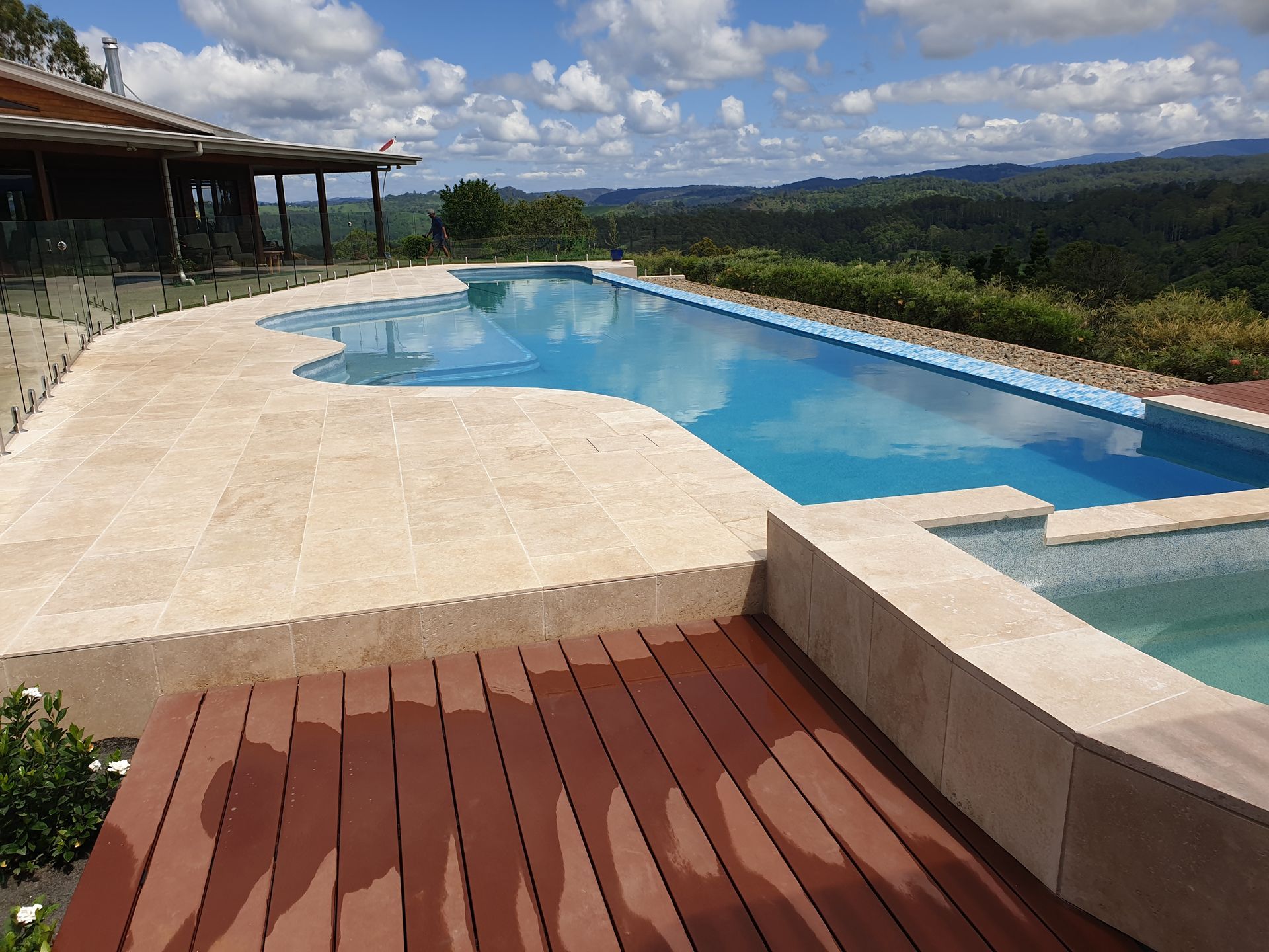 Swimming pool with blue water, tan stone patio, and wooden deck overlooking a forested landscape under a blue sky.