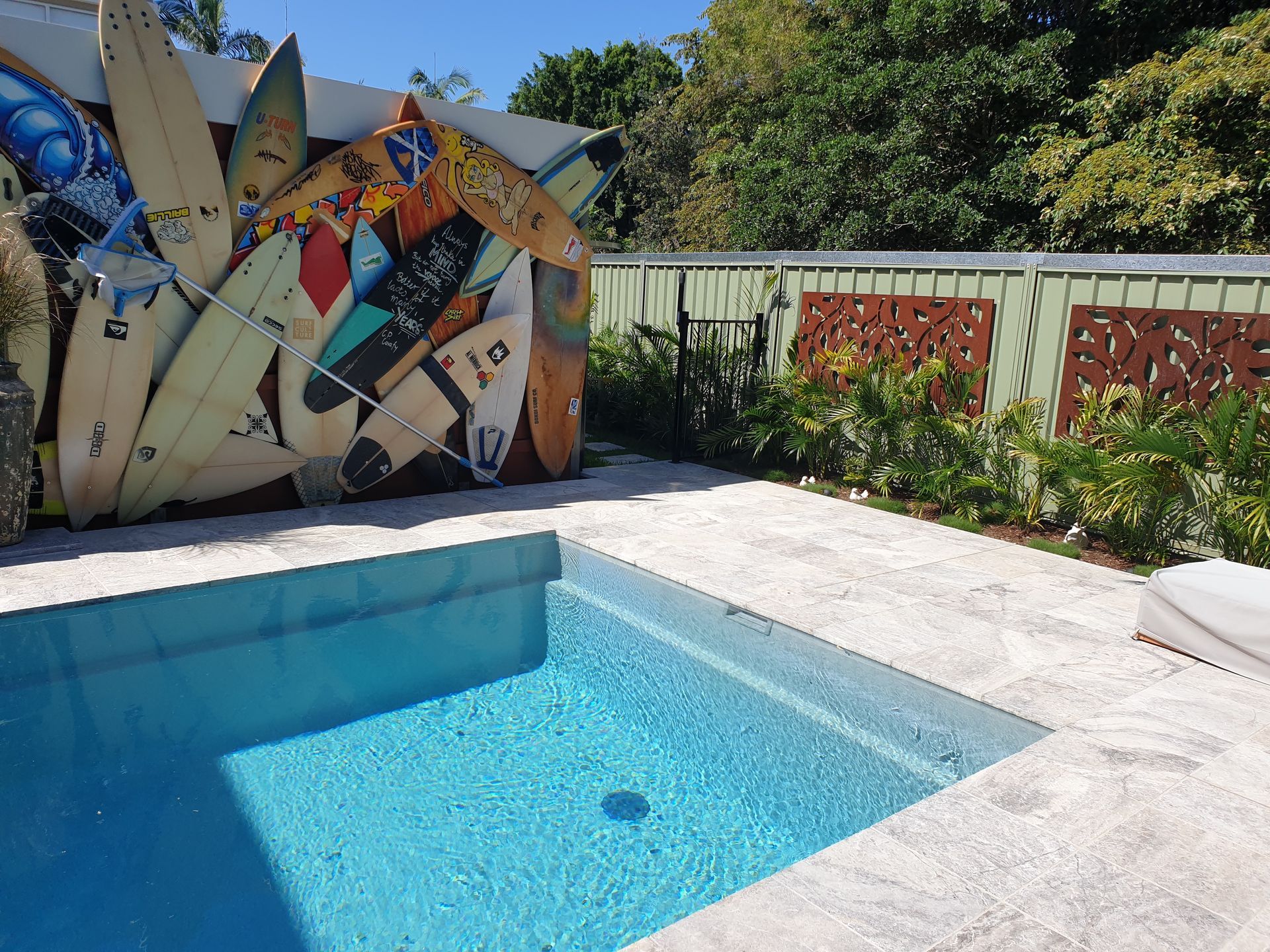 A small, blue swimming pool with a wall of surfboards next to a fenced yard.