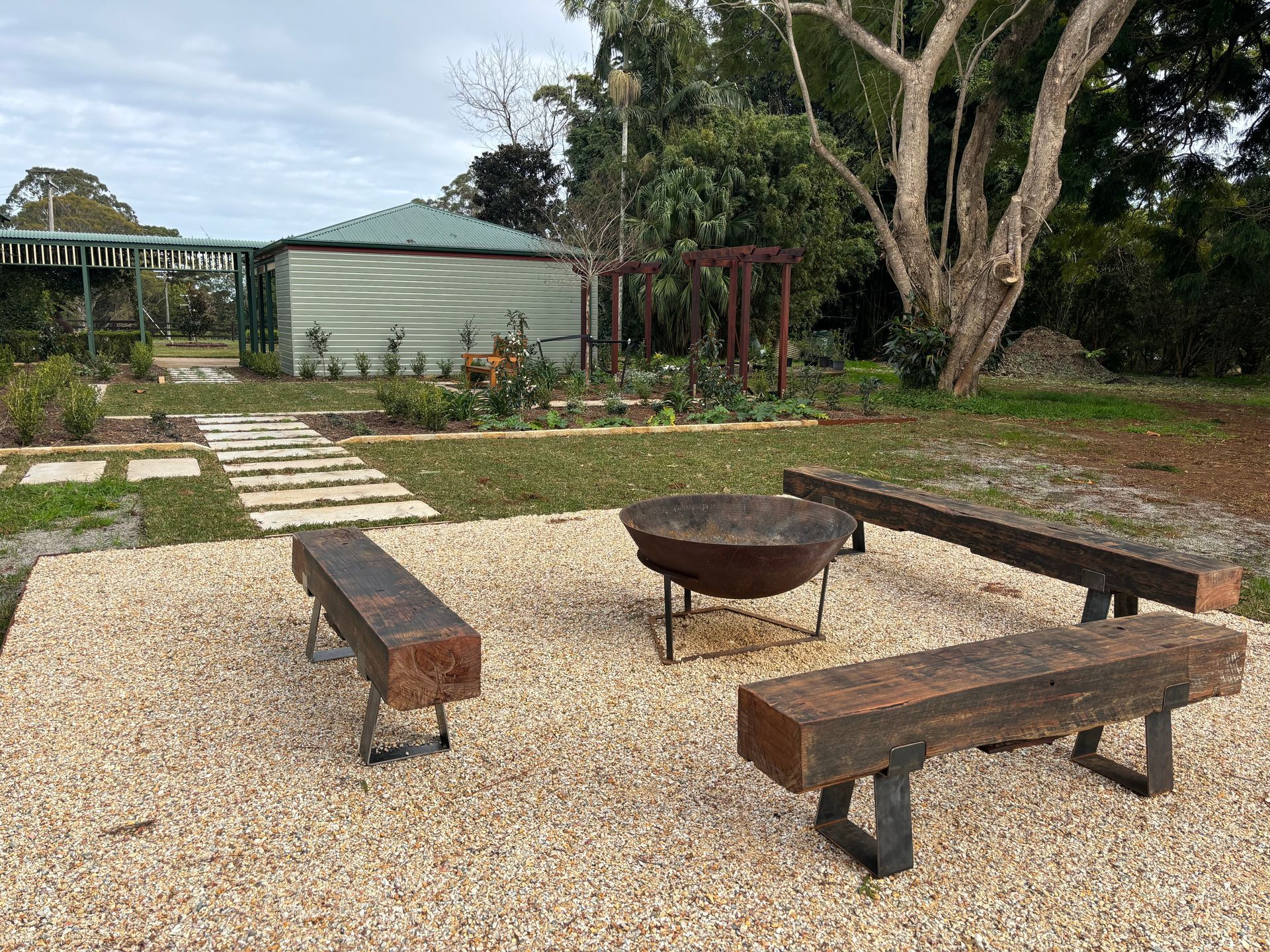 Backyard with gravel patio, fire pit, and wooden benches. Green structure and stone path in background.