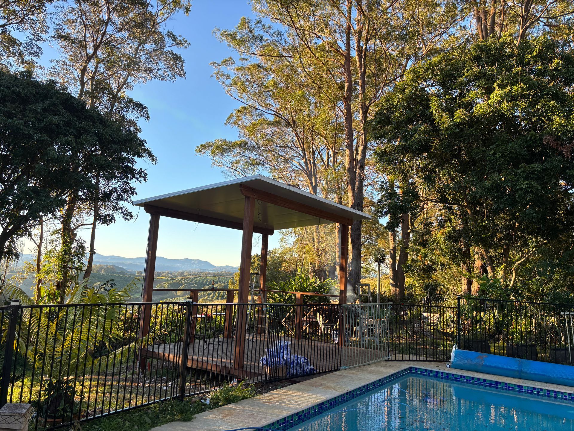 Poolside gazebo overlooking a valley, trees, blue sky, and black fence.