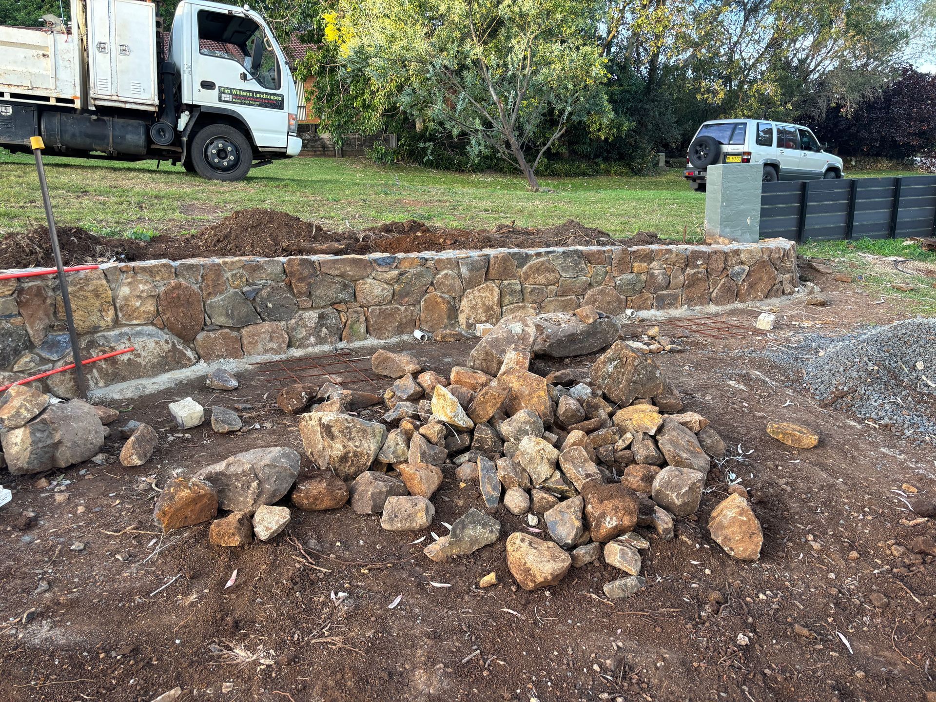 Stone retaining wall construction site, with a pile of rocks in foreground. Truck and car are in the background.