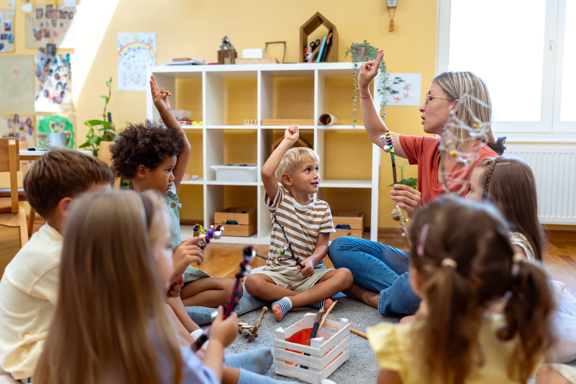 Teacher leading a circle of children in a classroom activity, all holding wands and raising their hands.