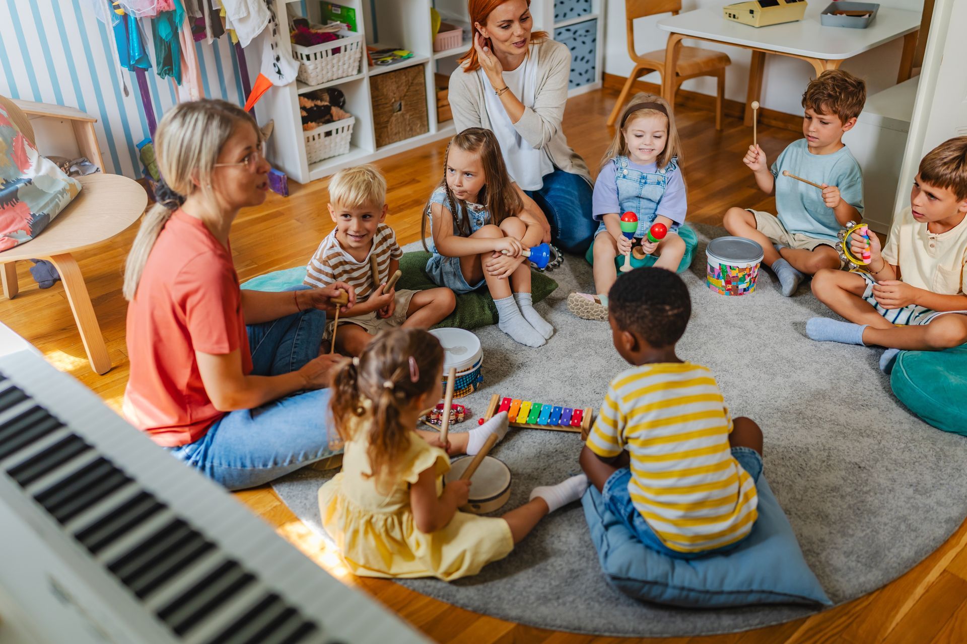 Children and two teachers in a music class, playing instruments while seated in a circle on the floor.