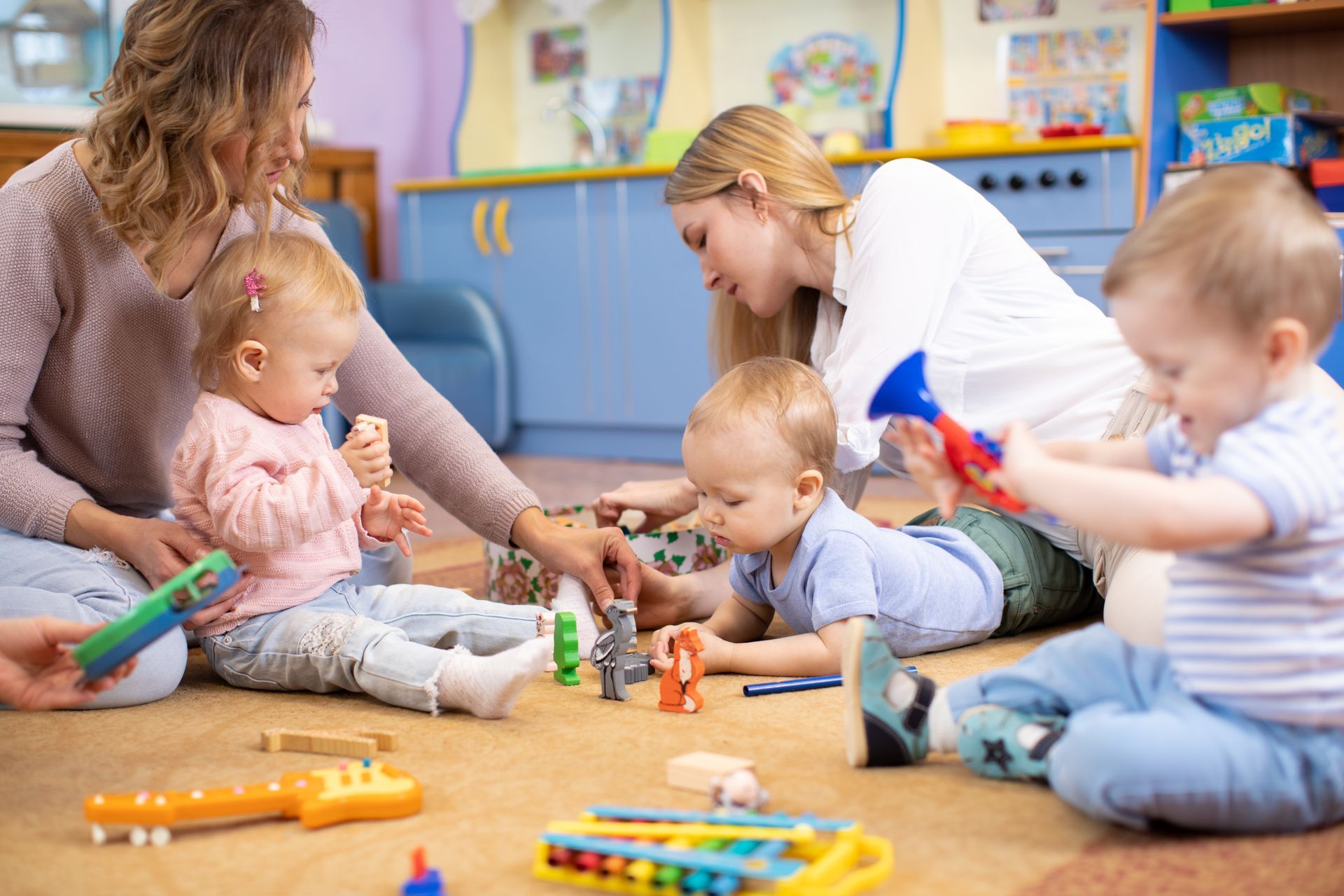Two women and three babies playing with toys on a playroom floor.