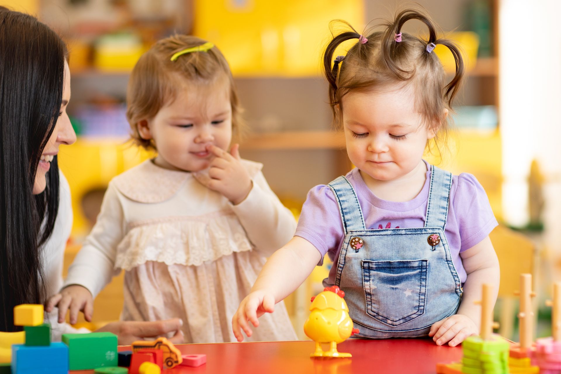 Teacher with two toddlers at a table playing with toys; one is touching her nose.