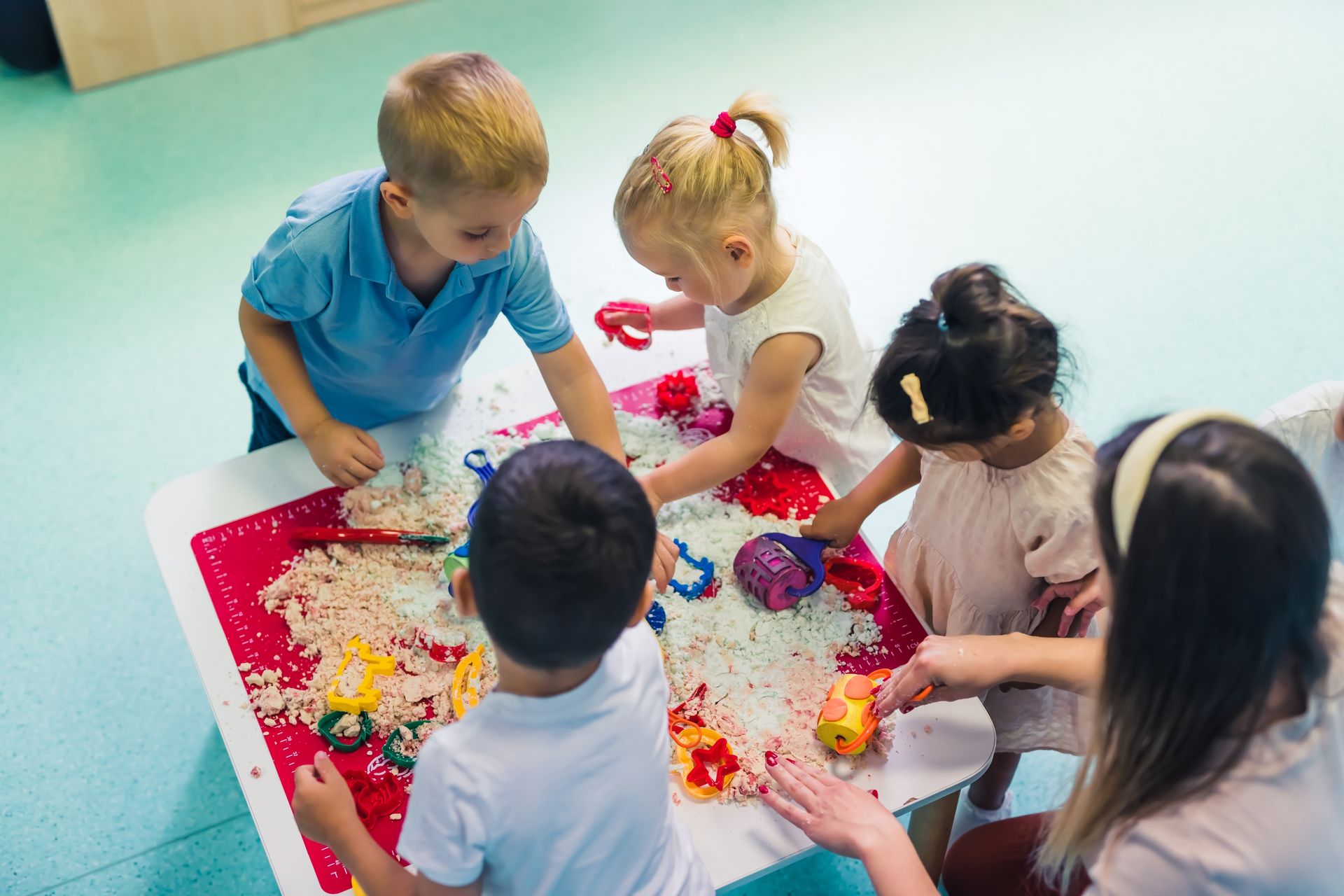 Children and an adult playing at a table with sensory materials; red tray, beige rice.