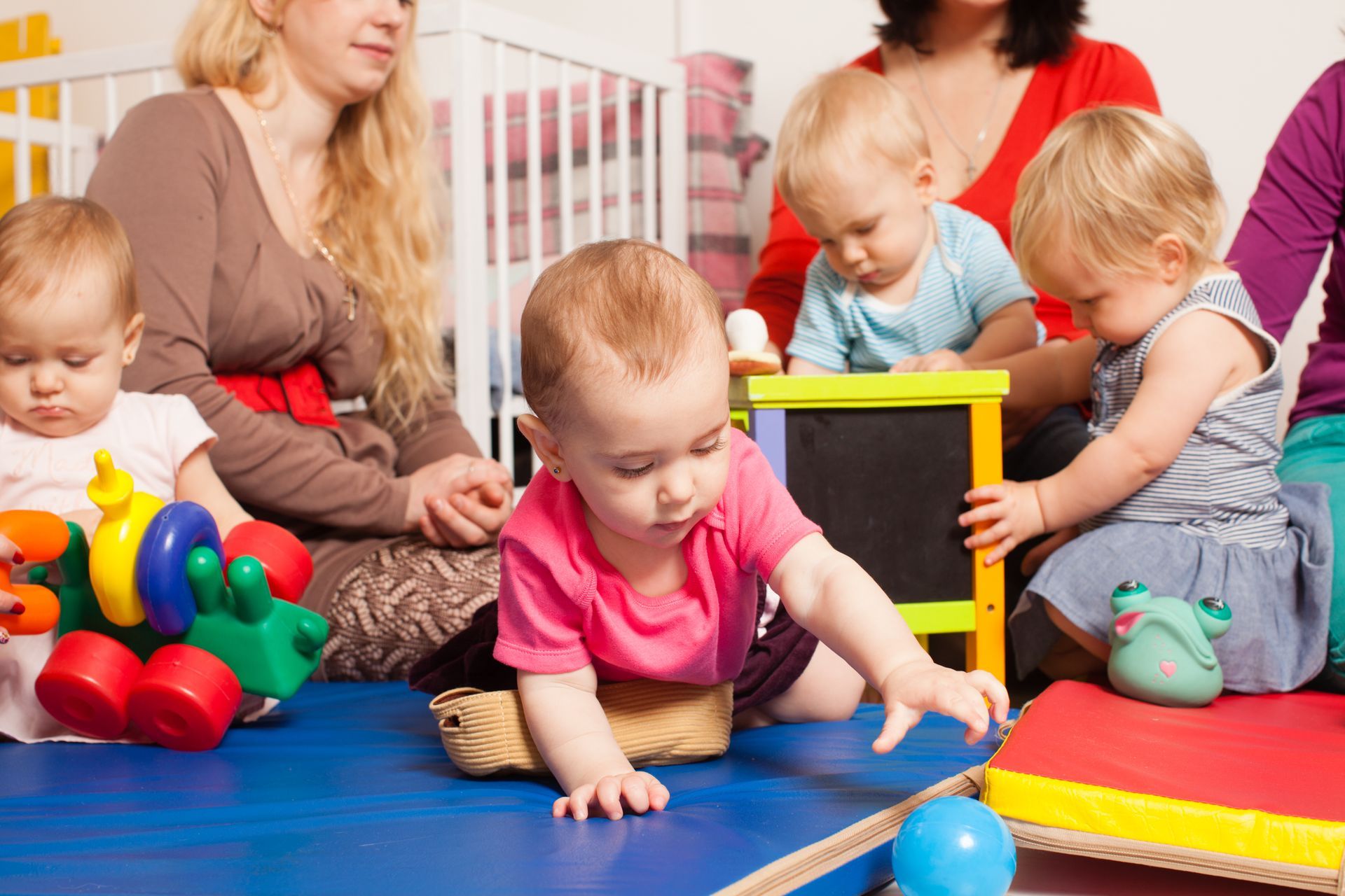 Babies crawl and play with toys on a blue mat with adults nearby.