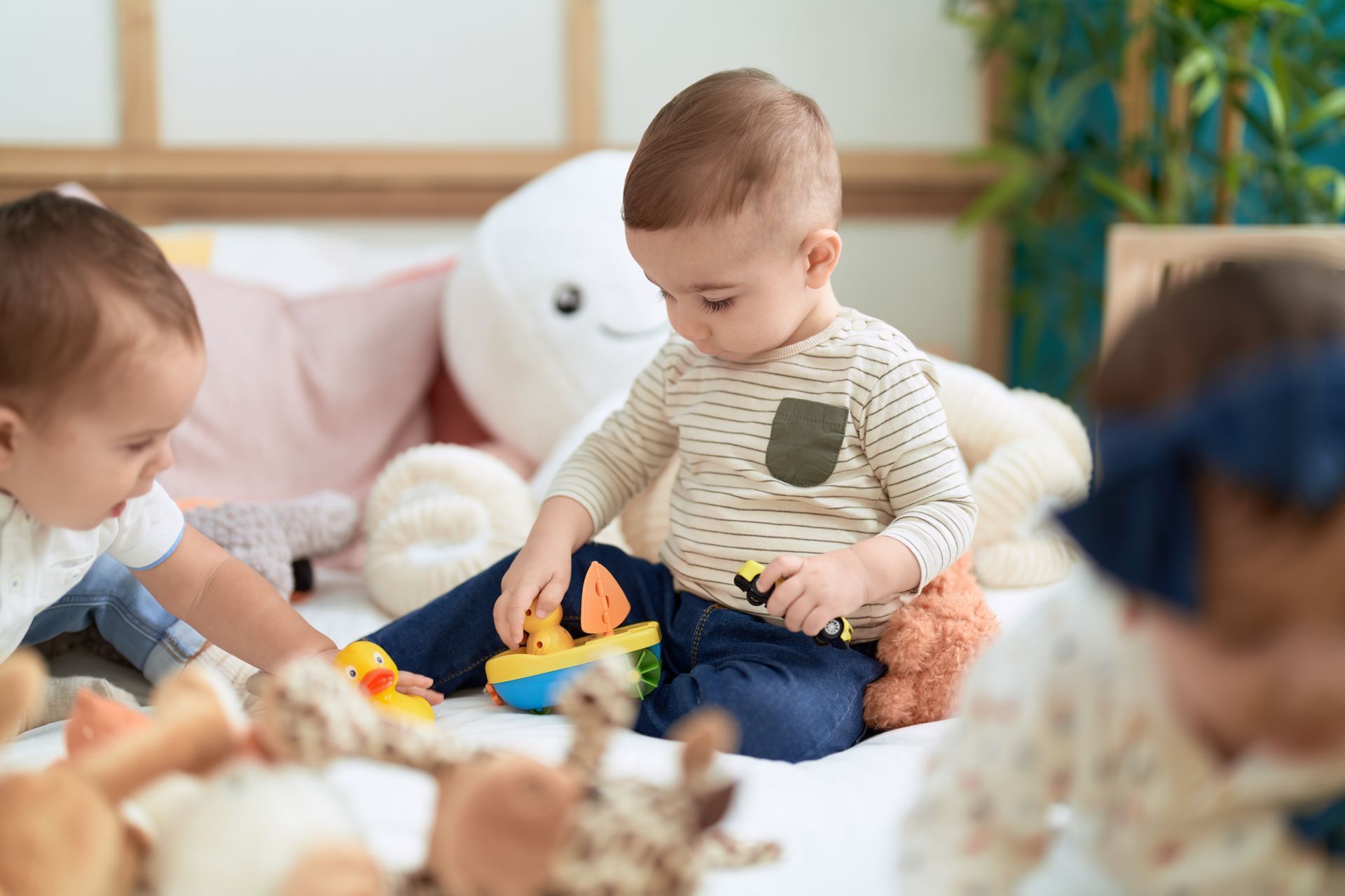 Three toddlers playing with toys on a bed surrounded by plush animals.