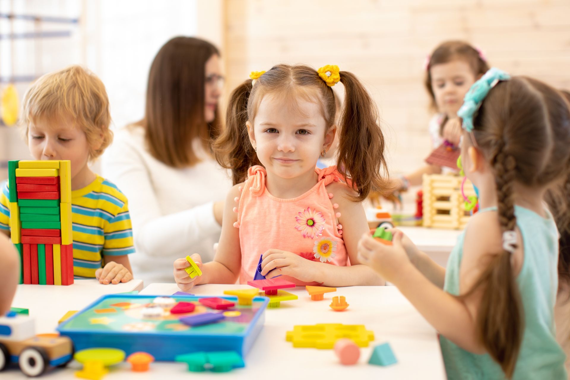 Children and teacher at a table playing with colorful blocks and toys in a brightly lit room.