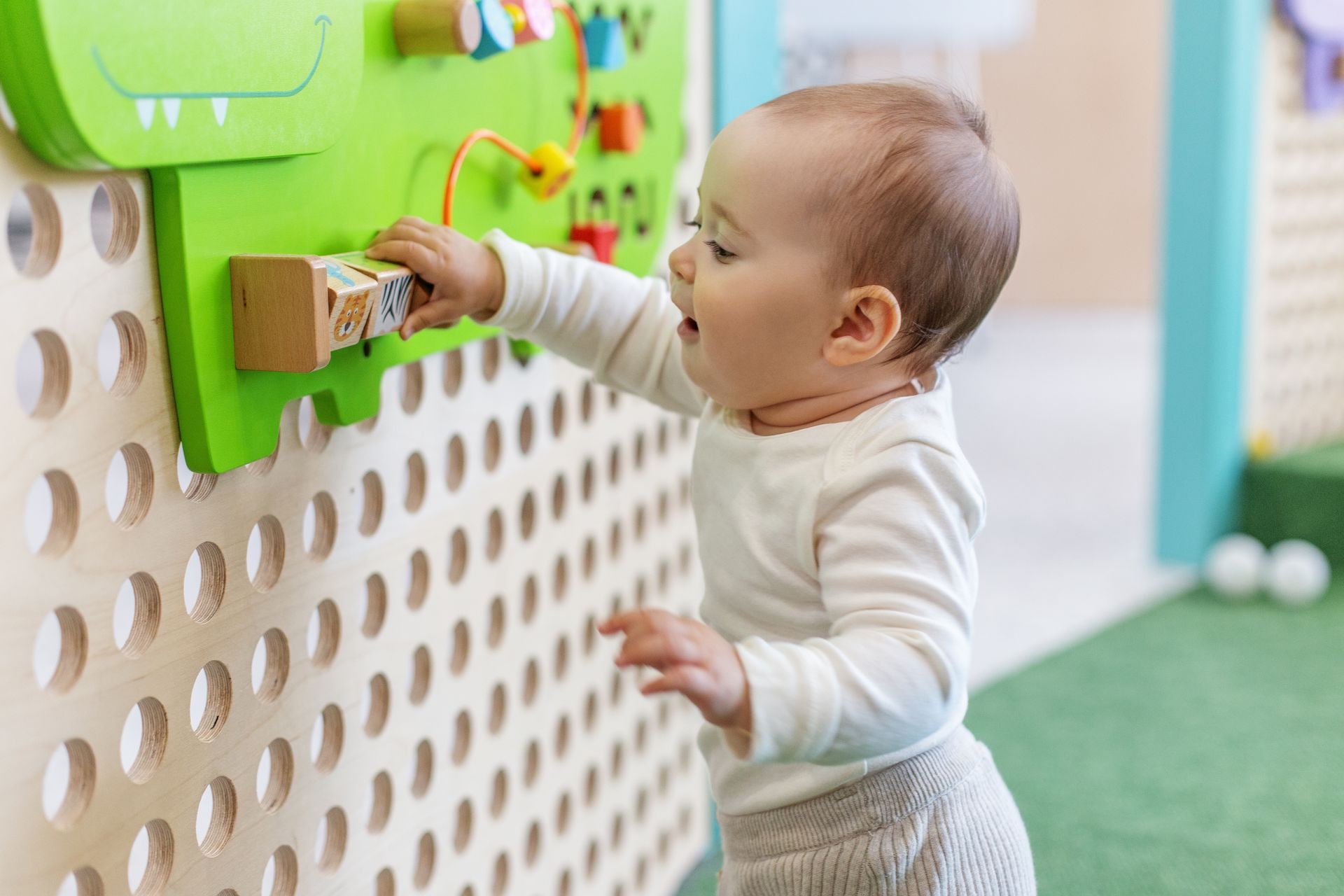 Baby reaching for a wooden block on a colorful sensory board with a focused expression.