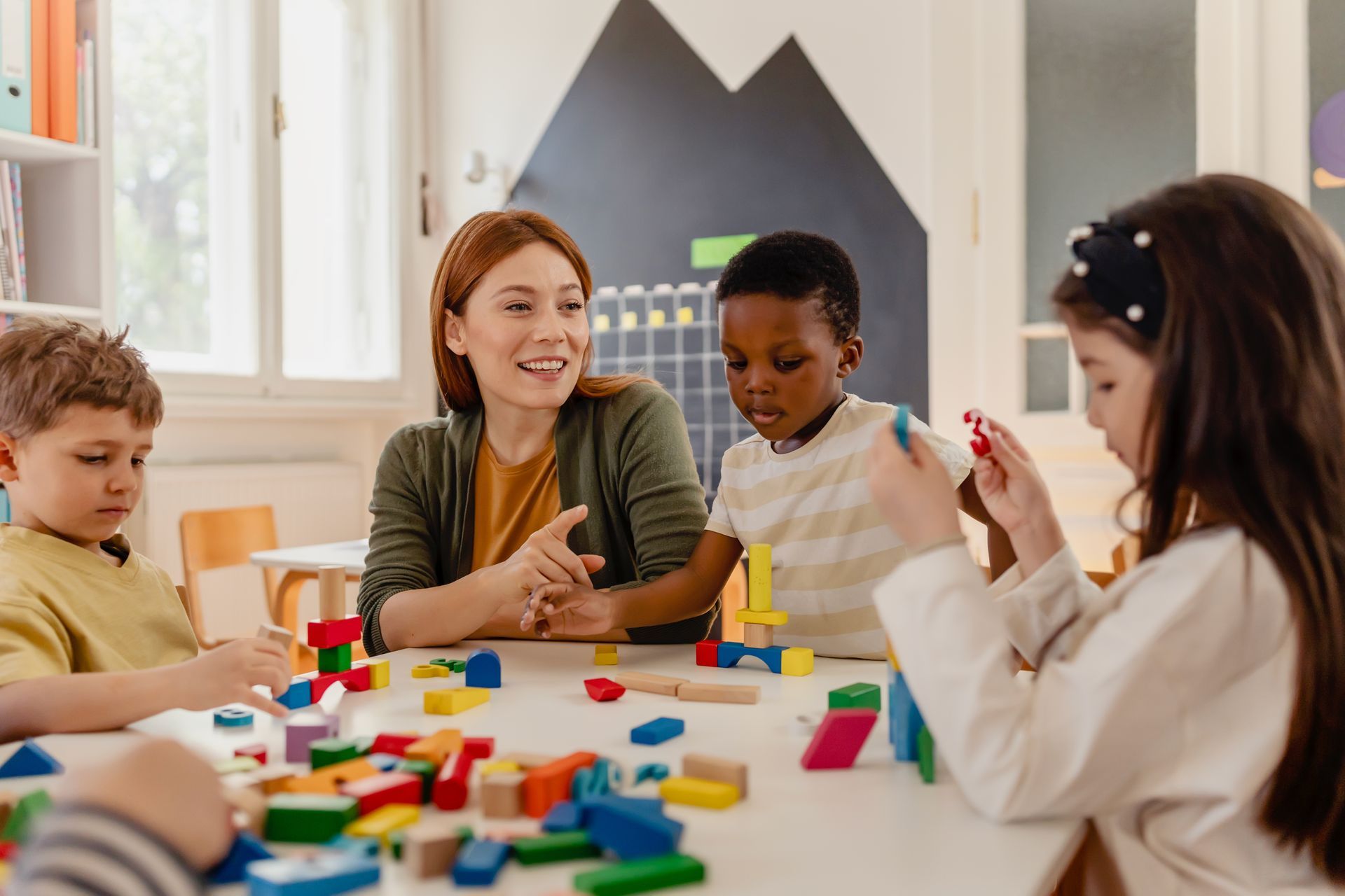 Teacher and children at a table playing with colorful blocks in a classroom.