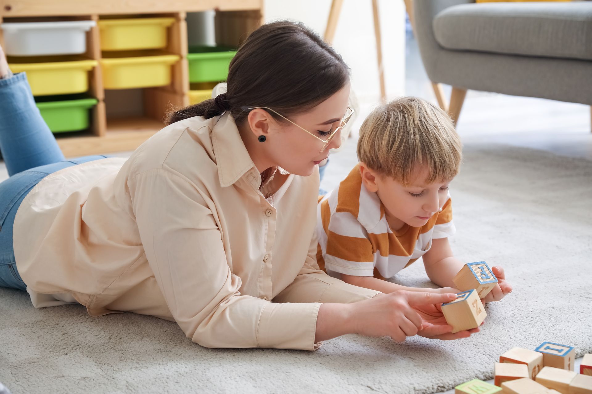 Woman and child playing with blocks on the floor. Beige top, glasses. Blocks are wood.