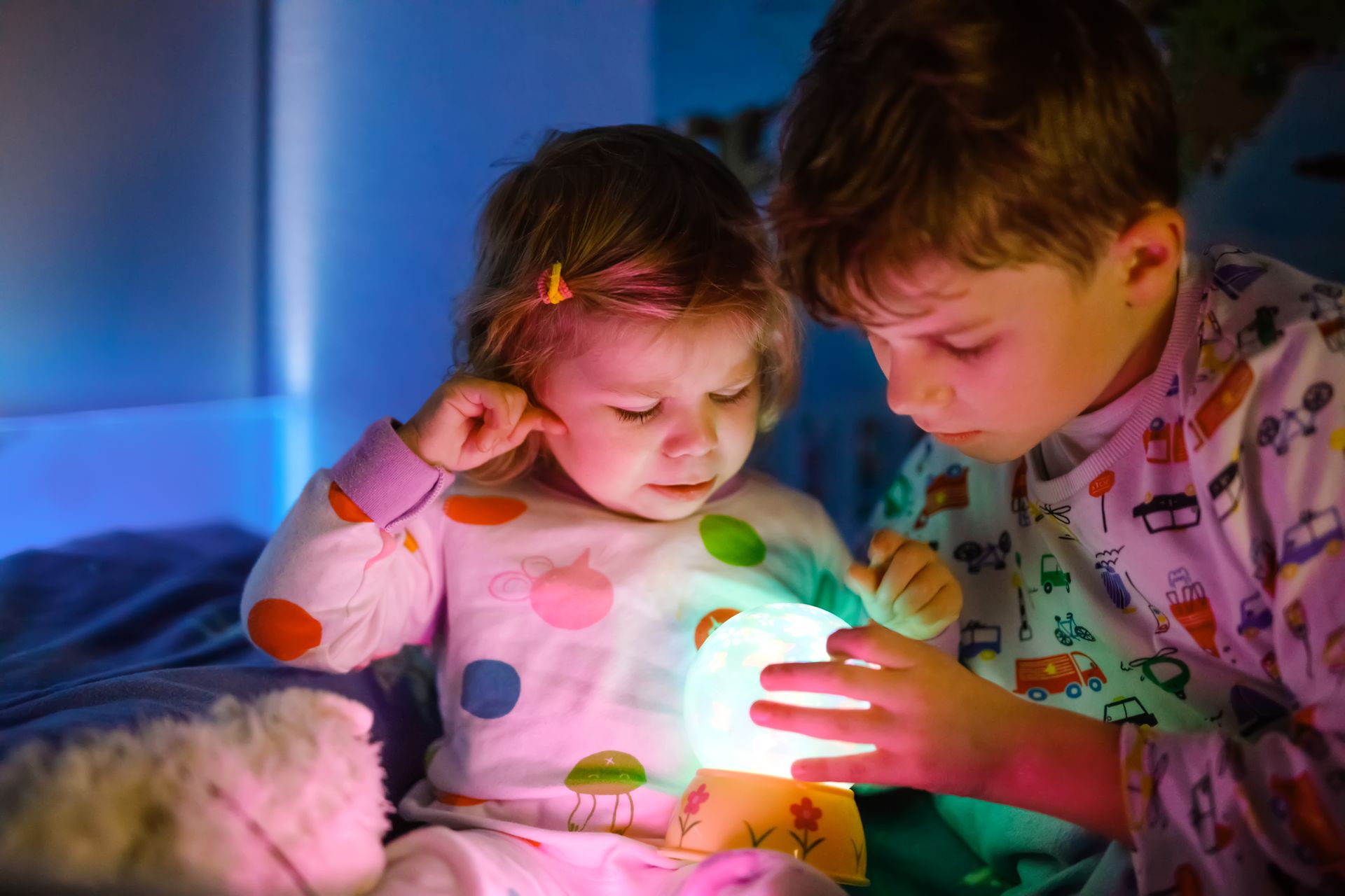 Two children in pajamas examine a glowing nightlight on a bed.