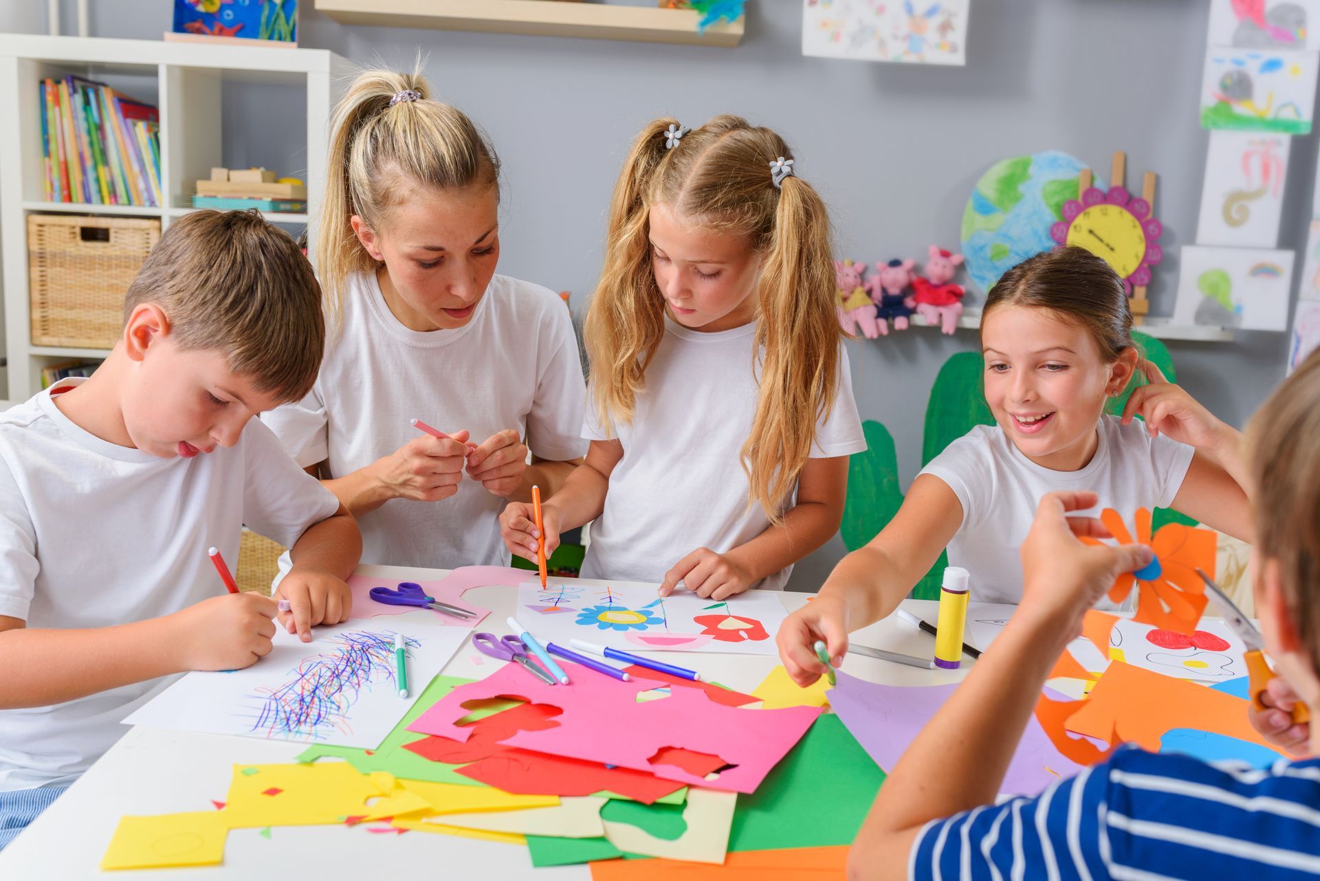 Children and an adult crafting at a table with colorful paper and art supplies in a classroom setting.