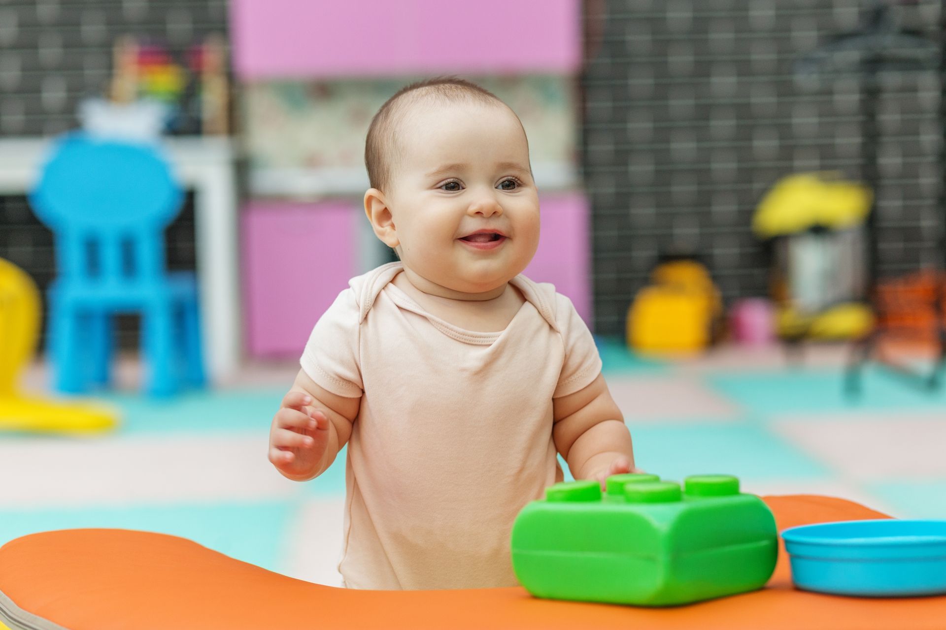 Smiling baby stands near a table with a green block and blue bowl in a colorful playroom.