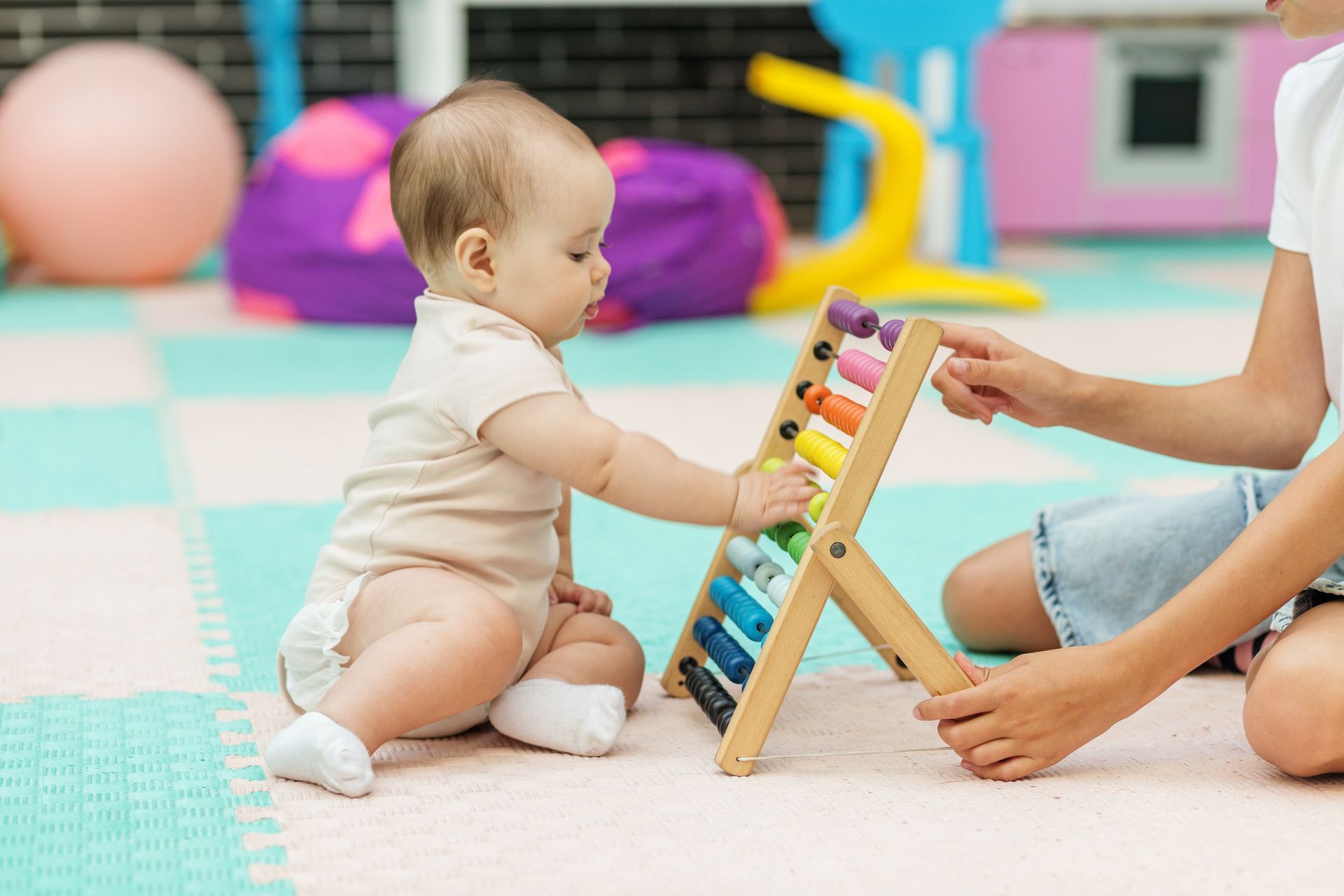 Baby playing with an abacus, sitting on a colorful mat with an adult.
