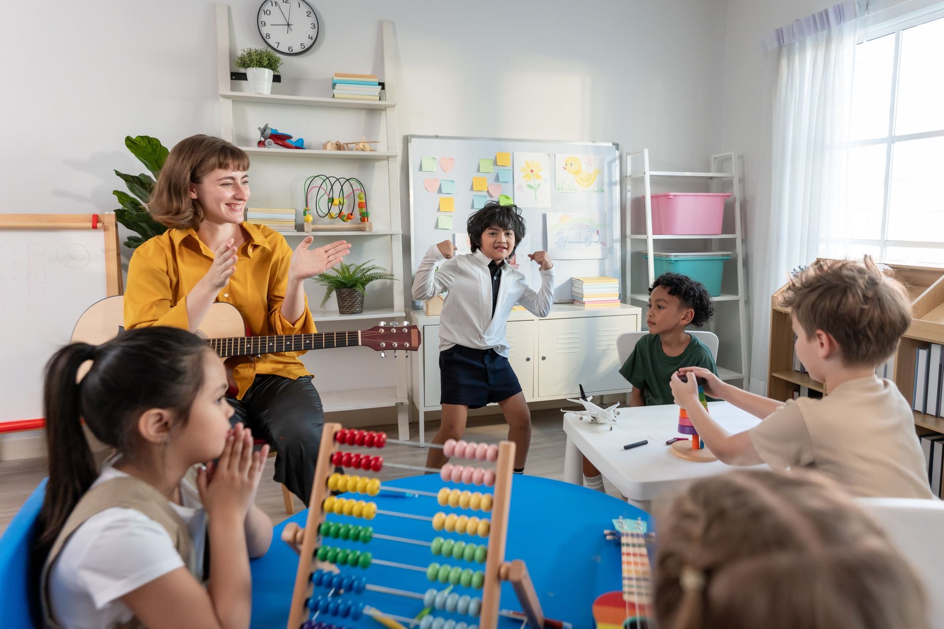 Teacher with guitar and children in a classroom; one child dances, others watch.